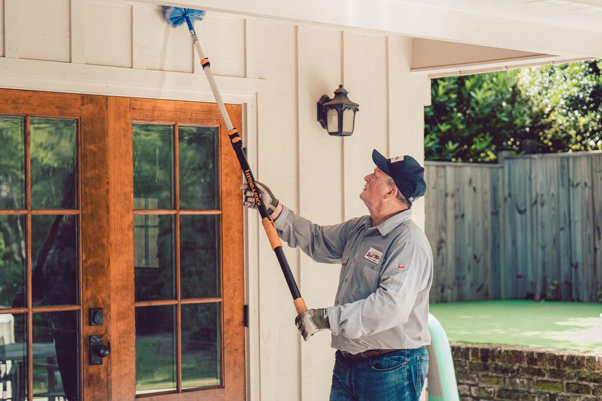 Man using extendable pole brush to clean home exterior soffit above wooden French doors
