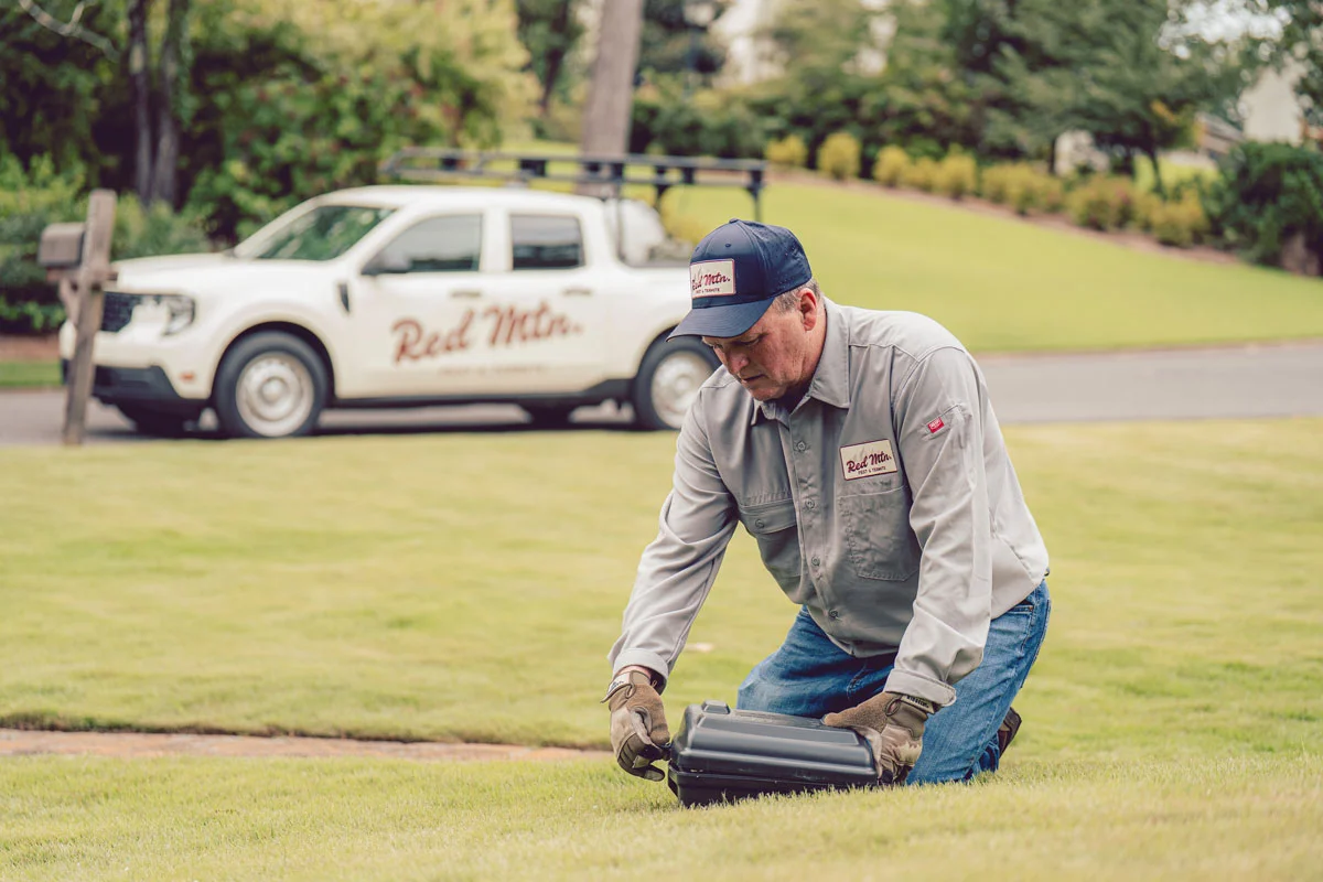 A man lawn technician kneeling on grass adjusting irrigation controller, company service truck in background