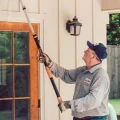 A man painting house exterior with an extension pole, touching up doorframe and window trim