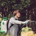 A man in gloves using backpack sprayer for pest control on garden shrubs near a brick wall
