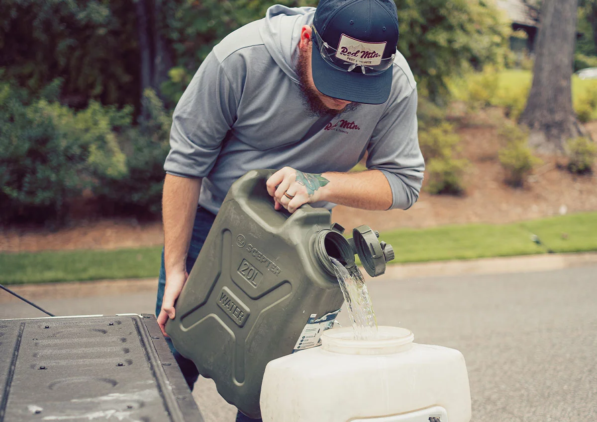 Man pouring water from 20L jerry can into a white container on a truck tailgate outdoors