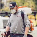 A man pest control technician wearing mask and cap operates backpack sprayer near service truck in suburban yard