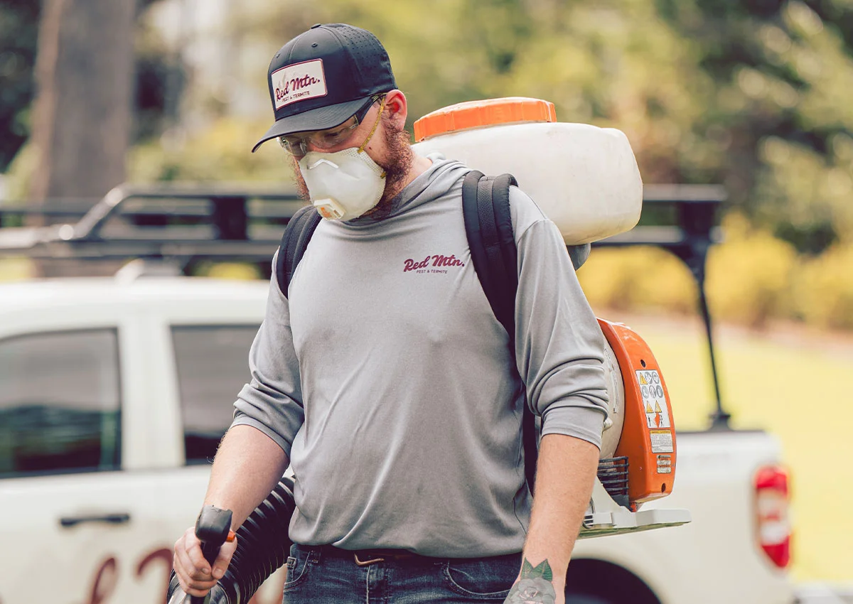 A man pest control technician wearing mask and cap operates backpack sprayer near service truck in suburban yard