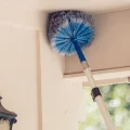 Blue round duster on telescopic pole cleaning soffit and eaves of beige house near outdoor lantern