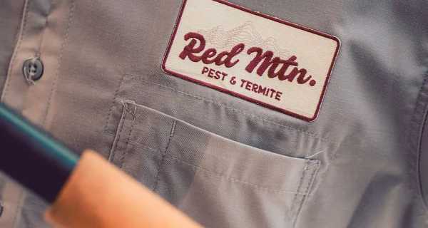Close-up of pest control uniform with Red Mtn. Pest & Termite embroidered patch on gray shirt and tool handle