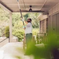 A man cleaning a covered patio ceiling with an extension pole on a residential porch, exterior maintenance