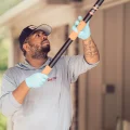 A man in a cap and gloves holding a telescoping pole inspecting house eaves for pest control