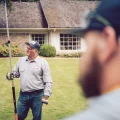 A man in work uniform holding a pole pruner on a suburban lawn in front of a brick house