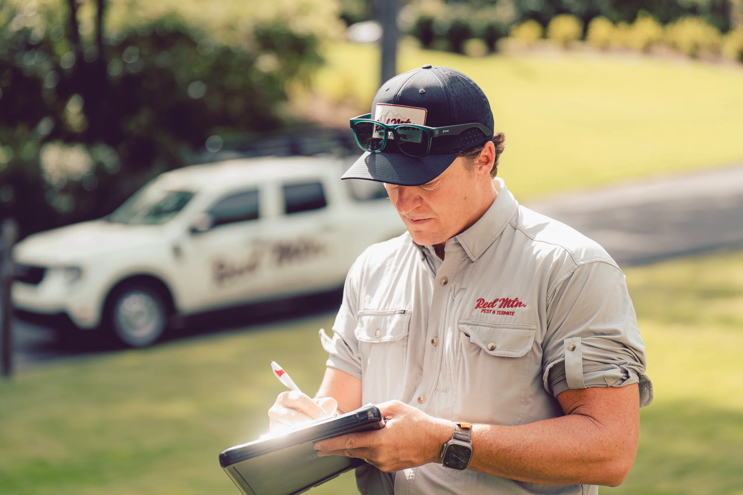A man pest control technician in uniform and cap writing on clipboard, company truck blurred in background
