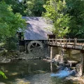 Historic rustic watermill with wooden water wheel and stone footbridge over a calm millpond surrounded by lush green trees.