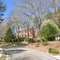 Large brick colonial home on curved, tree-lined suburban street with landscaped yard under clear blue sky